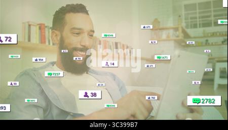 Homme barbu défilant tenant la tablette sur le canapé dans le salon moderne, avec des icônes de réaction flottantes Banque D'Images