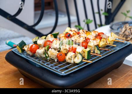 Une série de brochettes de poulet et de légumes maison grillant sur un barbecue électrique Moulinex, posé sur une table en bois à l'extérieur. Un di sain et coloré Banque D'Images