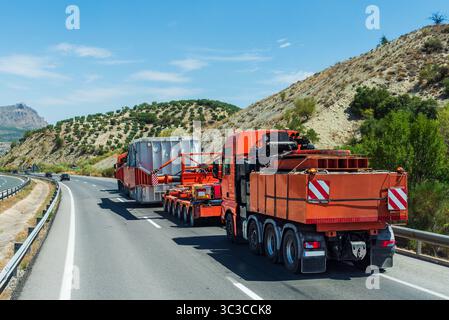 Camions lourds transportant une caisse métallique massive sur une pente raide, avec un camion devant et un autre derrière la charge. Vue arrière. Banque D'Images