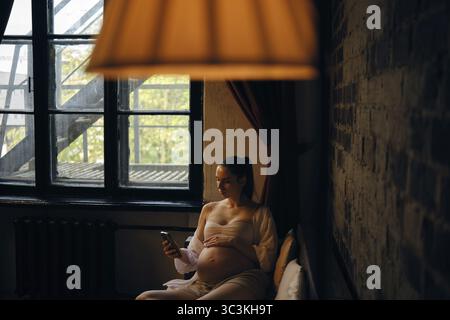 Dans une pièce confortable remplie de lumière naturelle, une femme enceinte s'assoit confortablement sur un canapé et fait l'ordre de livraison sur l'application mobile Banque D'Images