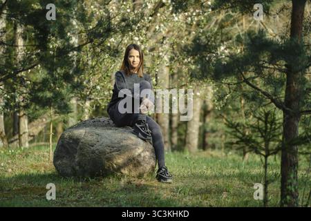 Une jeune femme est assise sur un grand rocher au milieu d'une forêt verte vibrante remplie d'arbres en fleurs. La lumière du soleil filtre à travers les arbres, cre Banque D'Images