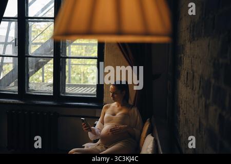 Une femme enceinte s'assoit confortablement dans sa maison confortable, berçant délicatement sa bosse de bébé tout en regardant son téléphone. La lumière naturelle filtre à travers le wi Banque D'Images