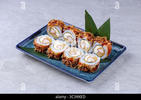 Rouleaux de sushi croustillants fourrés de crevettes, de fromage à la crème et d'oeuf, garnis d'oignons frits et de sauce sucrée, servis sur une assiette en céramique bleue. Banque D'Images