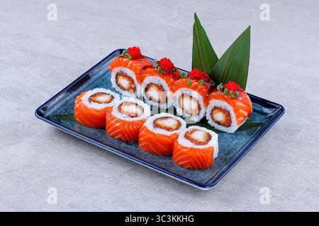 Rouleaux de sushi enveloppés de saumon et fourrés de crevettes tempura, recouverts de tobiko rouge et de microgreens, servis sur une assiette décorative bleue. Banque D'Images