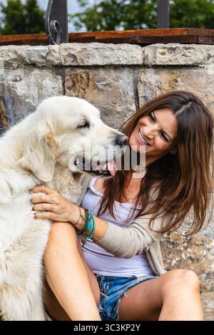 Femme joyeuse dans la tenue d'été décontractée embrasse avec amour son retriever d'or tout en étant assise sur l'herbe à côté d'un mur de pierre rustique, partageant une maman chaude et candide Banque D'Images