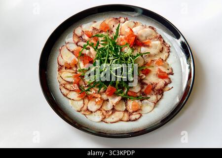 Une assiette magnifiquement agencée avec un carpaccio de poisson cru tendre garni d'Arugula fraîche et de tomates coupées en dés, mettant en valeur l'élégance culinaire et la fraîcheur Banque D'Images