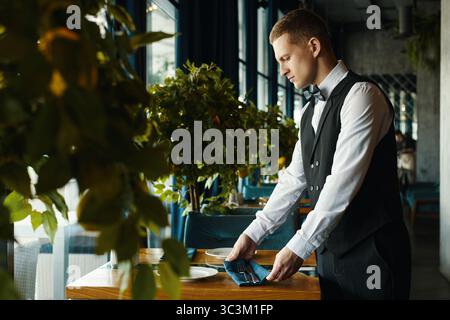 Jeune homme adulte caucasien organisant la table dans le restaurant, debout à côté d'une table en bois avec des assiettes et des couverts, portant un gilet formel et un noeud papillon, se préparant pour les invités Banque D'Images