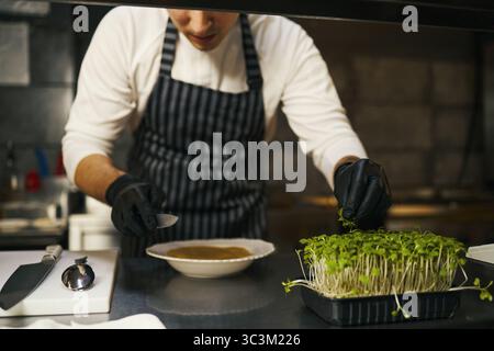 Jeune homme caucasien adulte portant un tablier rayé et des gants noirs préparant un plat gastronomique dans une cuisine professionnelle, organisant des microgreens frais sur une assiette avec une attention concentrée Banque D'Images