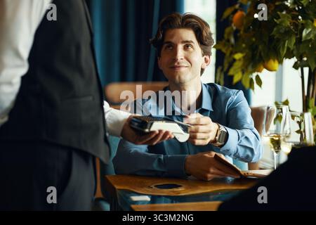 Jeune homme caucasien adulte assis à la table du restaurant remettant la carte de crédit au serveur tenant le terminal de paiement, souriant et faisant un contact visuel, verres à vin et arbre en arrière-plan Banque D'Images