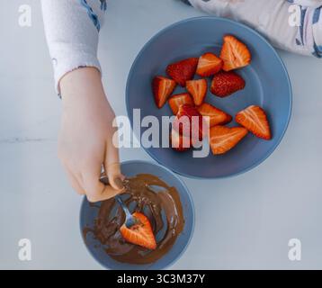Vue de dessus d'une main d'enfant trempant une fraise fraîche dans du chocolat fondu avec une fourchette. Bol à proximité avec des fraises tranchées. Banque D'Images