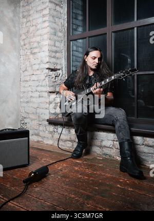 Jeune musicien de rock (headbanger) jouant de la guitare électrique. Portrait d'un bel homme aux cheveux longs. Prise de vue en intérieur, pleine longueur Banque D'Images