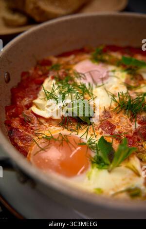 Un délicieux plat copieux de Shakshuka avec des œufs pochés dans une sauce tomate épicée garnie d'herbes fraîches et accompagné de pain chaud Banque D'Images