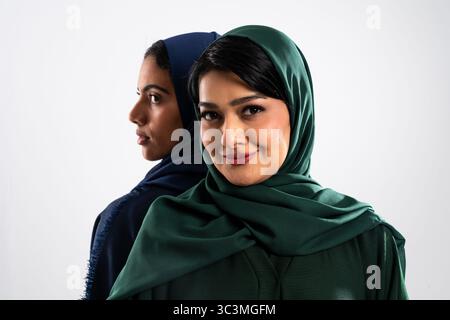 Deux femmes d'affaires musulmanes portant un hijab posent ensemble, dos à dos, dans un studio avec un fond blanc propre, à l'air confiant et élégant Banque D'Images