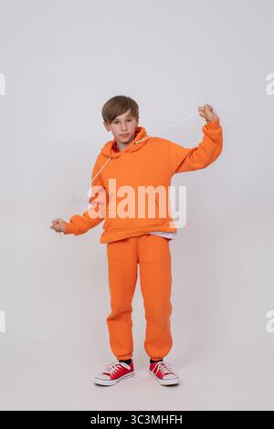 Un garçon dans un sweat-shirt orange et un pantalon orange tire les ficelles de sa capuche et regarde l'appareil photo. Le garçon porte des baskets rouges. Banque D'Images