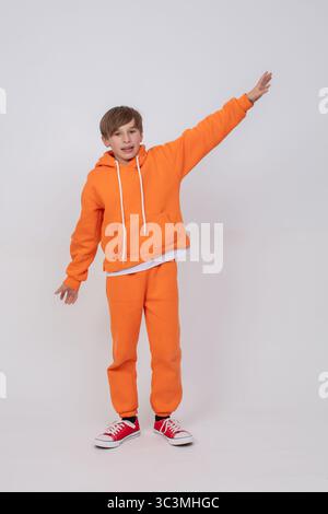 Un garçon portant un sweat-shirt orange et un pantalon orange leva la main et regarda la caméra. Le garçon porte des baskets rouges. Banque D'Images