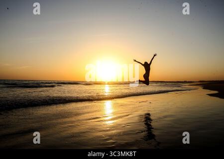 Silhouette de femme sportive en faisant un saut spectaculaire avec les bras levés sur la plage devant un beau coucher de soleil doré sur la mer au Portugal. Banque D'Images