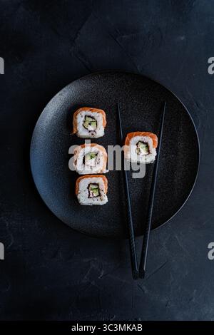 Vue de dessus du rouleau de sushi californien enveloppé dans du saumon, élégamment plaqué sur une plaque de céramique noire texturée avec des baguettes noires, serti contre un mat foncé Banque D'Images