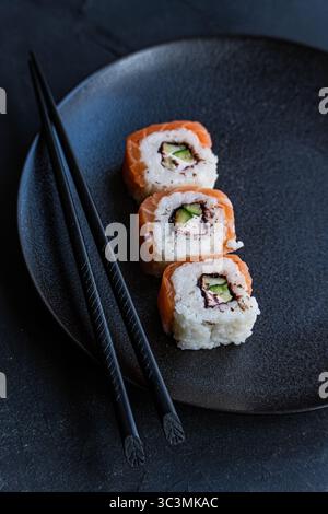 Trois rouleaux californiens recouverts de saumon sont assis sur une plaque de céramique foncée, accompagnés de baguettes noires. Le poisson vibrant et les ingrédients frais créent Banque D'Images