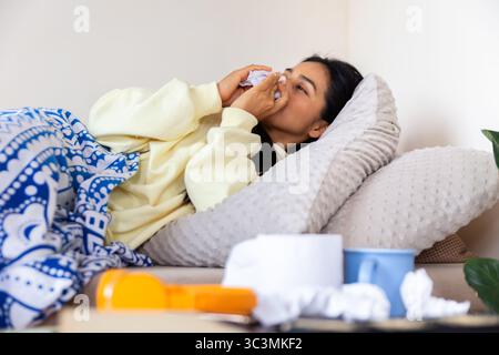 Une femme latine est allongée dans son lit à la maison, paraissant malheureuse et utilisant des mouchoirs pour se réconforter. Elle est enveloppée dans une couverture chaude, mettant l'accent sur le repos pendant elle Banque D'Images