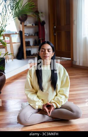 Une femme latine médite paisiblement à la maison, assise les jambes croisées sur le sol dans une pièce ensoleillée. L'environnement serein reflète sa tranquillité intérieure Banque D'Images