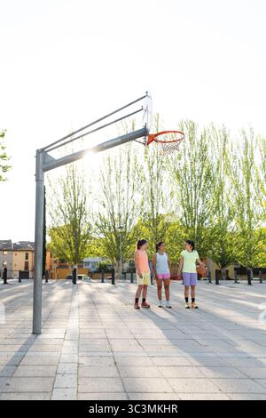 Trois femmes participent à un match de basket-ball de rue animé sur un terrain extérieur. Le soleil brille à travers les arbres, créant une atmosphère vibrante et énergique i Banque D'Images
