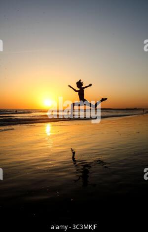 Silhouette de femme sportive en faisant un saut spectaculaire avec les jambes et les bras séparés sur la plage en face d'un beau coucher de soleil doré sur la mer. Banque D'Images
