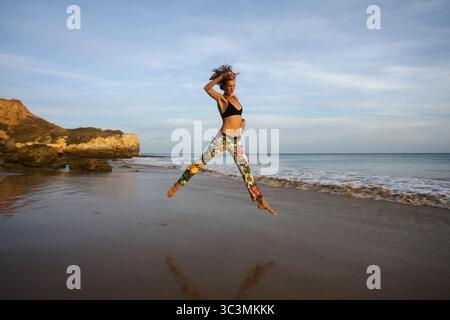 Jeune femme sportive en faisant un saut spectaculaire avec les jambes et les bras écartés sur la plage en face de la formation de rochers et de la mer au Portugal. Banque D'Images