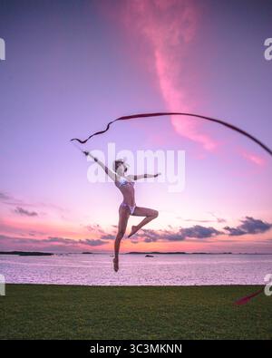 Femme sportive en bikini faisant un saut de danse avec ruban pour la gymnastique rythmique à la main devant un beau coucher de soleil sur la mer. Mode de vie actif Banque D'Images
