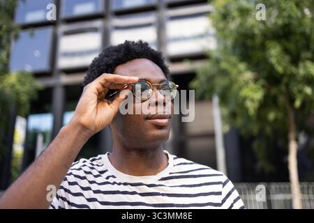 Un élève ajuste ses lunettes tout en regardant au loin, exsudant confiance et concentration. La toile de fond urbaine complète son style décontracté, suggesti Banque D'Images