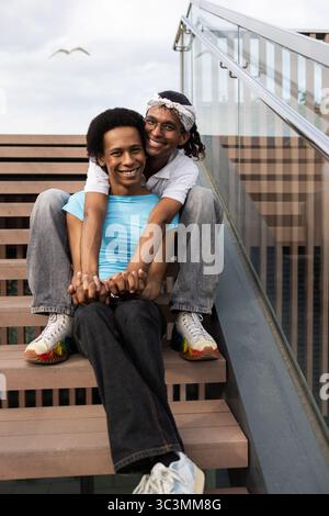 Un joyeux couple queer assis sur des escaliers extérieurs, profitant d'une journée d'été ensoleillée. Ils embrassent et sourient brillamment, capturant un moment de bonheur Banque D'Images