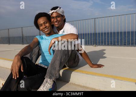 Un joyeux couple queer est assis ensemble un jour ensoleillé au bord de la mer. Ils sont souriants et apprécient le temps chaud de l'été, avec un océan calme dans le bac Banque D'Images
