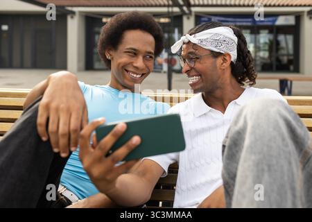 Un joyeux couple queer profite d'une journée ensoleillée ensemble, souriant et partageant un moment léger sur un banc. Leur bonheur est contagieux dans ce vibran Banque D'Images