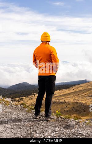 Un randonneur solitaire en équipement orange vif se dresse sur un sentier de montagne, contemplant le vaste paysage en contrebas. La scène suggère l'aventure, l'exploration, et Banque D'Images