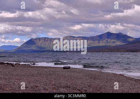 Le rivage de galets rencontre le lac McDonald vitreux sous les nuages couvrants tandis que les pics boisés du parc national des glaciers s'étendent sur un horizon tranquille. Banque D'Images