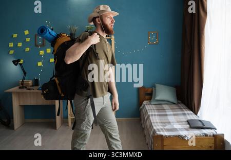 Portrait de randonneur barbu avec sac à dos se préparant pour une randonnée debout dans une pièce à la maison, vue de côté Banque D'Images