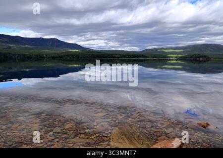 Les eaux claires et peu profondes révèlent des pierres colorées du lit du lac tandis que les nuages miroirs et les collines forestières ensoleillées dansent sur le lac McDonald dans le parc national des glaciers. Banque D'Images
