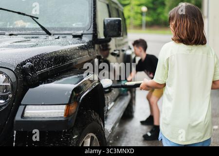 Deux frères et sœurs lavent activement un SUV noir avec de l'eau et des éponges dans une allée ensoleillée. Le cadre extérieur met en valeur le travail d'équipe et le plaisir pendant une voiture Banque D'Images