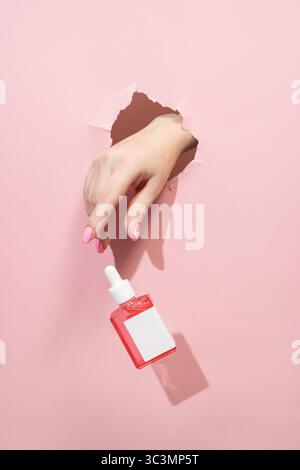 La main manucurée d'une femme émerge à travers un fond de papier rose déchiré, semblant lâcher une bouteille compte-gouttes de sérum rose, créant un style et moderne Banque D'Images