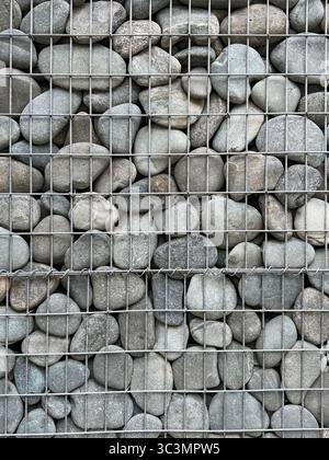 Vue rapprochée d'un mur de gabion rempli de roches lisses, illustrant le design industriel moderne. Le motif de grille crée une texture visuelle saisissante dans Banque D'Images