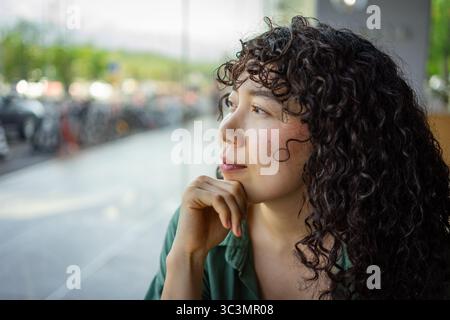 Une jeune femme aux cheveux bouclés regarde soigneusement à l'extérieur tout en étant assise à l'intérieur par une grande fenêtre. La lumière douce souligne son expression sereine et c Banque D'Images