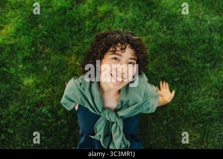 Une jeune femme joyeuse aux cheveux bouclés sourit brillamment tout en étant assise sur l'herbe verte dans un parc. Elle respire le bonheur et la détente dans un setti en plein air Banque D'Images