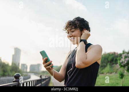 Une jeune femme aux cheveux bouclés sourit alors qu'elle aime la musique sur son smartphone tout en marchant à l'extérieur. Elle porte des écouteurs, exsudant la joie et la détente dans un lus Banque D'Images