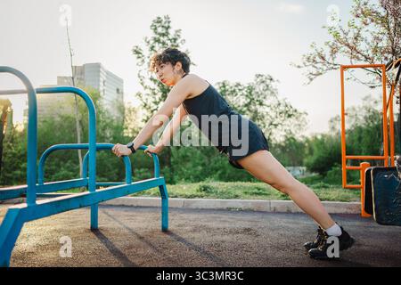 Une jeune femme aux cheveux bouclés s'engage dans un entraînement push-up en plein air, en utilisant un équipement de gymnastique bleu vif. Le cadre ensoleillé met en valeur sa forme athlétique et Banque D'Images