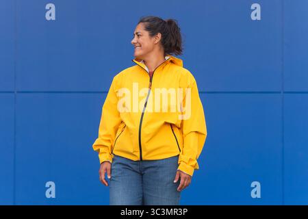 Femme portant un imperméable jaune vif se tient souriant devant un mur bleu vif. Son Jean denim décontracté complète la toile de fond colorée, Creati Banque D'Images