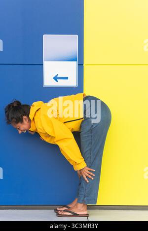 Une femme dans un imperméable jaune s'appuie contre un mur bleu et jaune vif avec un signe de flèche. Sa pose ludique crée un contraste vif avec l'audacieux Banque D'Images