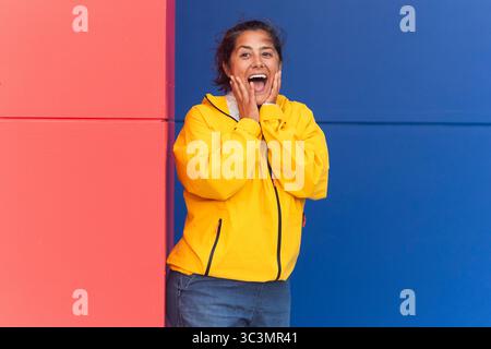 Une femme joyeuse portant un imperméable jaune éclatant se dresse sur un fond saisissant de murs rouges et bleus. Son expression respire l'excitation et le happ Banque D'Images