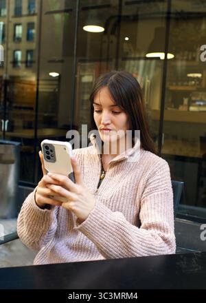 Une jeune femme est assise sur une terrasse concentrée sur son smartphone. Elle porte un pull douillet, alliant détente et connectivité dans un cadre extérieur moderne Banque D'Images