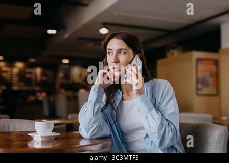 Une femme professionnelle s'engage dans un appel téléphonique alors qu'elle est assise dans un café confortable, mettant en valeur un mélange de travail et de détente. Une tasse de café est posée sur la table Banque D'Images