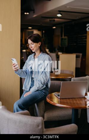 Une femme est assise dans un café confortable, engrossée dans son smartphone tout en travaillant. Une tenue décontractée et un ordinateur portable ouvert sur la table suggèrent un décontracté, productif à Banque D'Images