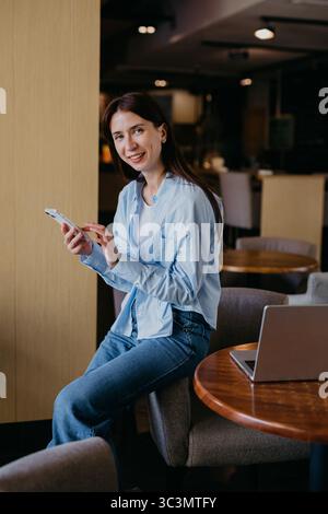 Une femme s'assoit confortablement dans un café confortable, s'engageant avec son smartphone et son ordinateur portable. Elle sourit et semble détendue, profitant de son environnement de travail, pendant ce temps Banque D'Images